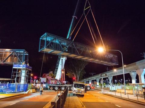 An on the ground view showing a crane lifting a segment of the pedestrian walkway  into place  at night at Sydney Metro's Kellyville Station.