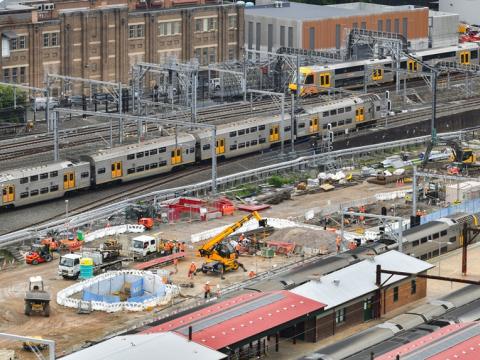A bird's eye view looking across at the construction site of the Sydney Metro station box at Central Station with many heavy machinery on the ground between the platforms as two trains are leaving the platform in the background.  