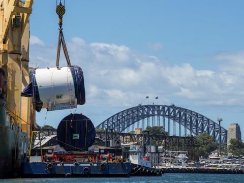 An on the ground view looking across at Tunnel Boring Machine being lifted off the boat by a large crane onto a smaller boat as it is arriving at Sydney Metro's The Bays construction site with shots of the Sydney Harbour Bridge in the background. 