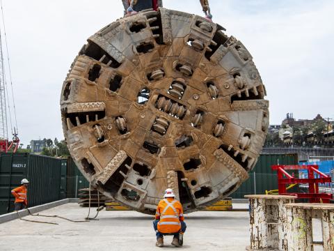 The cutterhead of tunnel boring machine Mum Shirl being lifted from the ground into the air by crane, whilst a construction worker supervises.
