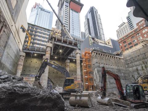 An on ground view looking up at heavy vehicle machinery working inside the Pitt Street South construction site. Tall buildings can be seen in the background.