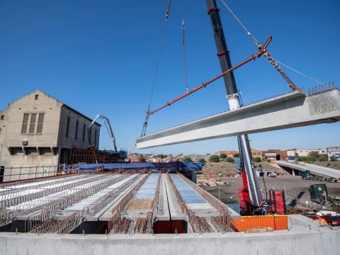 A crane lifting a beam into place at the Sydenham Station and Junction site. There is a building and blue sky in the background.