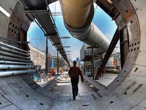 An on the ground view of a construction worker walking through Tunnel Boring Machine (TBM) 2 Florence with a large pipe overhead and construction site in the background. 