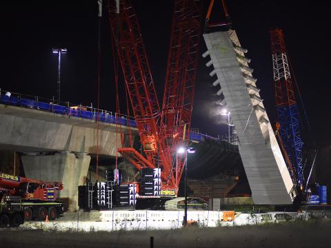 An on the ground view looking at the construction of the skytrain bridge tower as a crane lift is lifting a section of the bridge tower from the ground at night at Sydney Metro's Rouse Hill. 