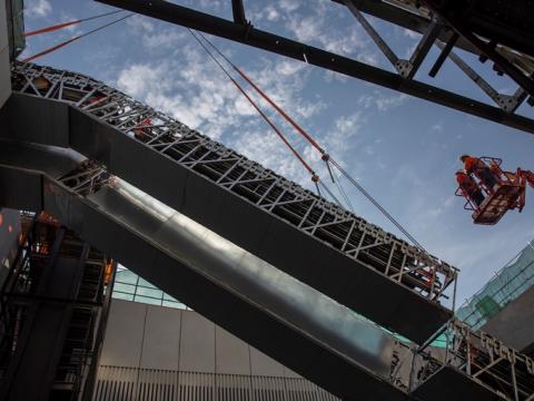 An on the ground view looking up as the double escalators are being crane lifted into place and two construction workers on a crane lift are inspecting the installation at Sydney Metro's Norwest site n May 2018. 