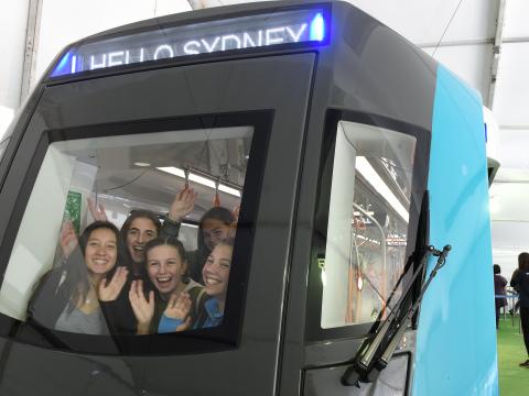 A group of five girls smiling and waving from the front of the Sydney Metro train carriage that was displayed at the Royal Easter Show. 