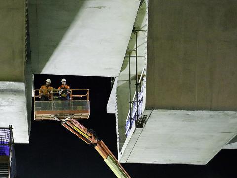 An on the ground view looking up at two construction workers on a crane lift as the final segment is nearing completion. 