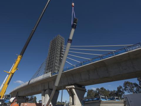 An on the ground view looking up at the construction of the railway bridge as cable poles are being lifted into place by a crane with the bridge tower in the background.