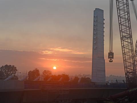 Looking across to the construction of Sydney Metro's skytrain bridge at sunrise. The image depicts a crane in the foreground with the bridge tower in the background. 