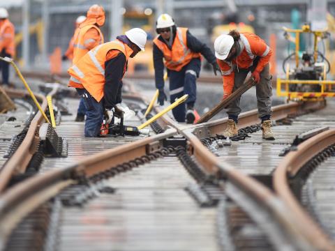A group of four construction workers using machinery to lay the train tracks at a cross section, with more constructions completing the same task in the background. 