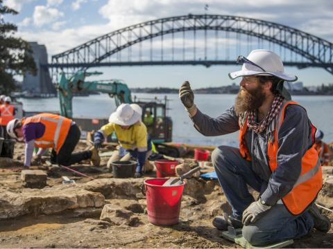 Archaeologists in high vis and hard hats are on their hands and knees while working in the excavation area at the Blues Point construction site. Sydney Harbour Bridge is seen in the background.