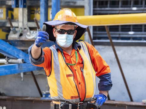A construction worker in orange high-vis, facemask and sunglasses gives the camera a thumbs-up while on site at Waterloo Station.
