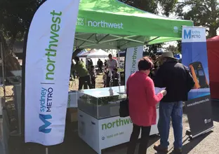 Community stall showing two community members in the Sydney Metro Northwest branded marquee looking at the Northwest station model, and reading a brochure. There are 2 branded tear drop banners and one branded Sydney Metro pull up banner. 