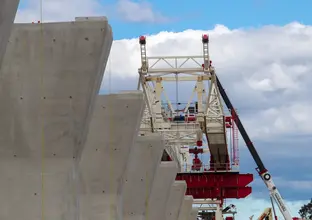 An on the ground view looking up at the gantry crane being lifted into place on top of raised concrete pillars for the construction of Sydney Metro's skytrain. 