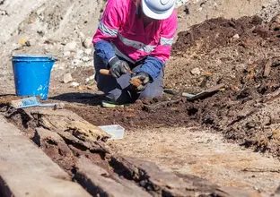 A female archaeologist in pink high vis and hard hat is on her hands and knees while working in the excavation area at the future site of Barangaroo Metro Station where an old boat was found.
