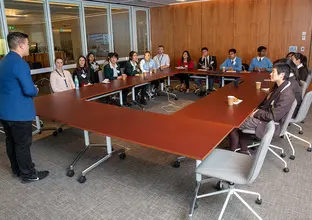 A man in a blue suit jacket is presenting to a several people sitting around an O-shaped table in a board room 