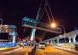 An on the ground view showing a crane lifting a segment of the pedestrian walkway into place at night at Sydney Metro's Kellyville Station.