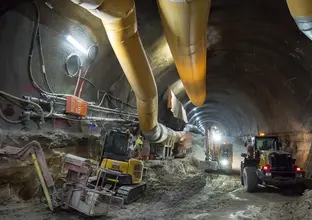 An on the ground view looking inside the tunnel at Sydney Metro's Pitt Street North construction site with two large pipes running through the cavern with a digger underneath he pipes and a Roadheader in the background.
