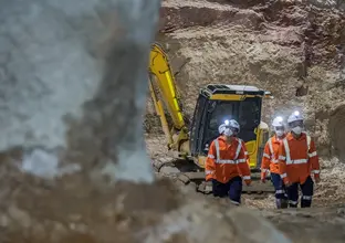 A close up shot of four construction workers walking inside a construction site with head torches shining bright inside with an excavator in the background at Sydney Metro's Martin Place Station.