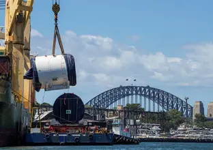 An on the ground view looking across at Tunnel Boring Machine being lifted off the boat by a large crane onto a smaller boat as it is arriving at Sydney Metro's The Bays construction site with shots of the Sydney Harbour Bridge in the background. 