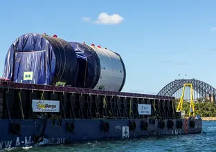 An on the ground view looking across at Tunnel Boring Machine on a boat as it is arriving at Sydney Metro's The Bays construction site with shots of the Sydney Harbour Bridge in the background. 
