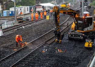 Machine moving the track whilst a construction worker cuts the track. There are several construction workers and shed in the background.