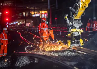 Machine cutting the train track at night. There is sparks coming off the track and 3 workers in personal protective equipment