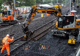 A heavy machine is picking up and moving the rail track. Construction worker in high viz is directing the operator of the machine.