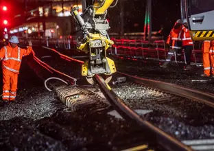 The arm of a heavy machine is picking up and moving the rail track at night time. Construction worker in high viz is directing the operator of the machine.