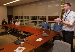 A man in a lanyard is presenting to a group of people are sitting around a U-shaped boardroom table.