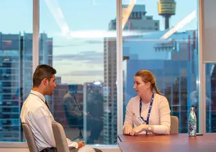 A man and woman are sitting down in discussion at a boardroom table with the city skyline in the background.