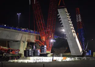 An on the ground view looking at the construction of the skytrain bridge tower as a crane lift is lifting a section of the bridge tower from the ground at night at Sydney Metro's Rouse Hill.
