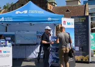 Close up shot showing the Sydney Metro community information stand with a branded marquee, alignment maps, brochures on display and pull up banners as a Sydney Metro employee is talking to a member of the community at Parramatta Farmers Markets.