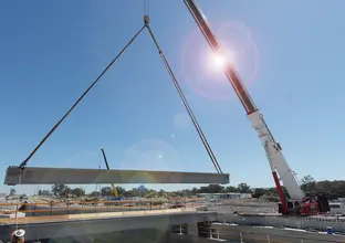 An on the ground view looking cross the construction of the bridge works as a crane is lifting a large piece of concrete material whilst a construction worker guides it into place with a rope at Sydney Metro's Kellyville Station. 