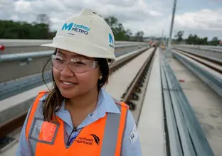 A woman in a Sydney Metro hard hat, safety glasses and high vis vest smiles while standing on the tracks.