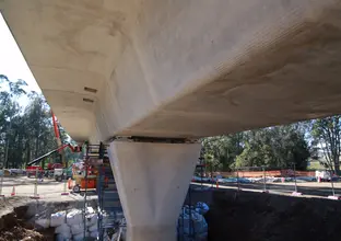 A close up view under the bridge as construction work continues on Sydney Metro's skytrain platform, there are three heavy machinery in the background behind a fence of the platform construction.