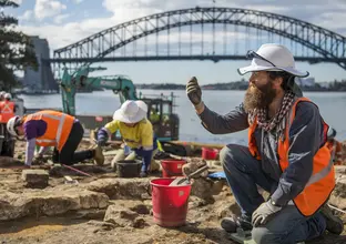 Archaeologists in high vis and hard hats are on their hands and knees while working in the excavation area at the Blues Point construction site. Sydney Harbour Bridge is seen in the background.