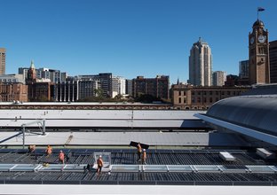 Wide shot of construction workers installing solar panels on Central Station's rooftop with Sydney's skyline in the background