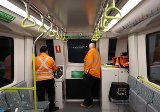 Two men in orange high-vis stand at the front of a metro train during testing at night.