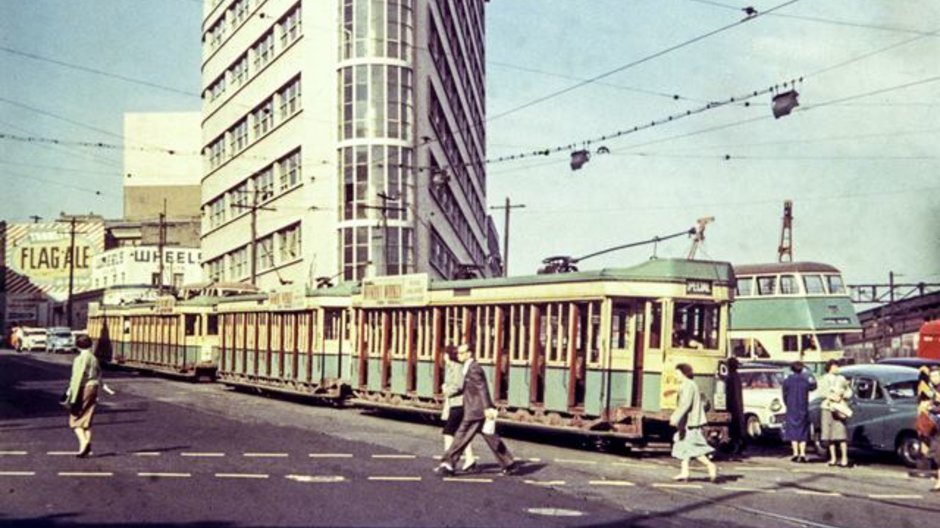 An image from 1957 of the corner of Elizabeth and Chalmers Street, Surry Hills showing commuters crossing the road with a tram on the tracks, cars parked and a bus in the background (Image is from City of Sydney Archives: 044664)