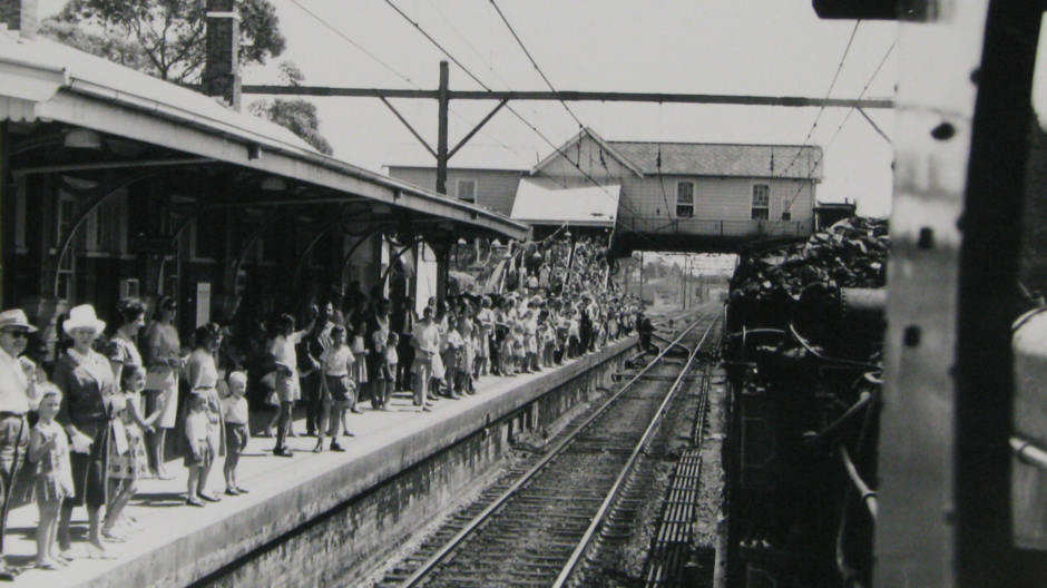 A black and white photo of commuters waiting at the platform as a steam train arrives at Chatswood Railway Station during Willoughby Centenary celebrations in 1965.