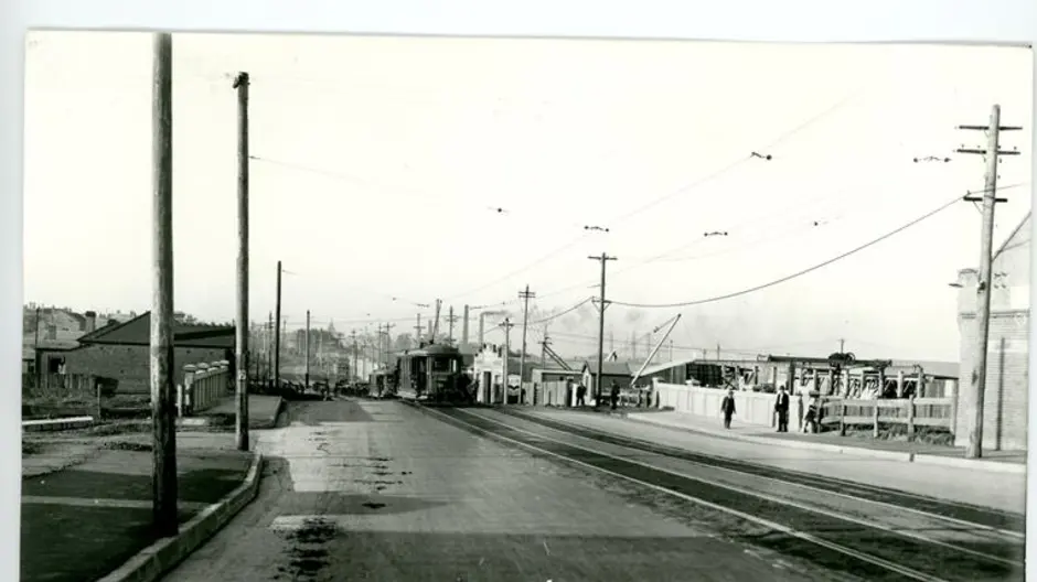 A black and white image of Cooks River Road, Sydnebgam from 1928 showing the road with old tram tracks to the right of the image and a tram in the background (Image Courtesy of Inner West Council Library and History)