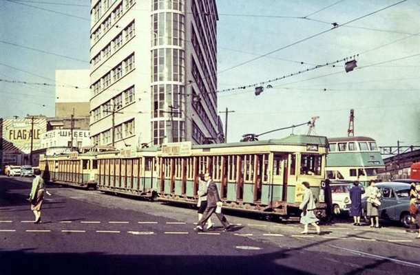 An image from 1957 of the corner of Elizabeth and Chalmers Street, Surry Hills showing commuters crossing the road with a tram on the tracks, cars parked and a bus in the background (Image is from City of Sydney Archives: 044664)