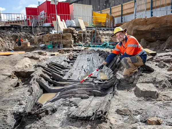 Man in orange high visibility shirt kneels down next to the remnants of boat found in Barangaroo.