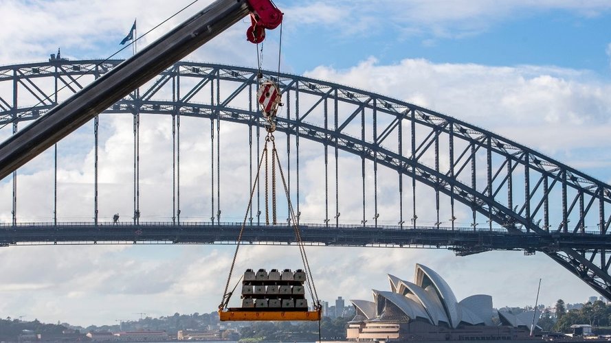 A crane is lowering sleepers for delivery to Blues Point constriction site. The Opera house and Harbour Bridge are in the background.