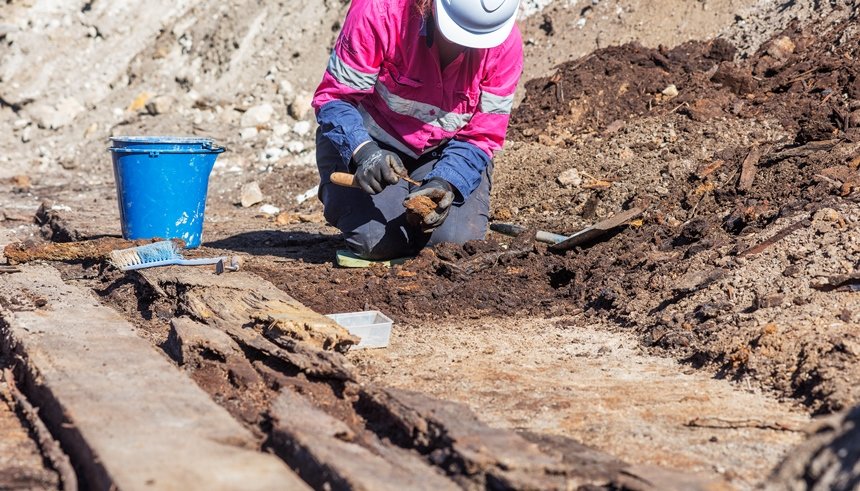 A female archaeologist in pink high vis and hard hat is on her hands and knees while working in the excavation area at the future site of Barangaroo Metro Station where an old boat was found.
