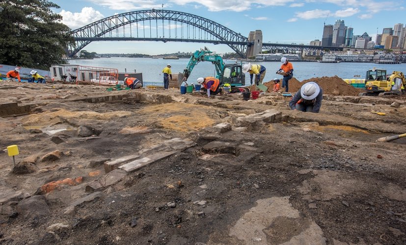 A wide angle of archaeologists in high vis and hard hats working in the excavation area at the Blues Point construction site. Sydney Harbour Bridge and city skyline is seen in the background.