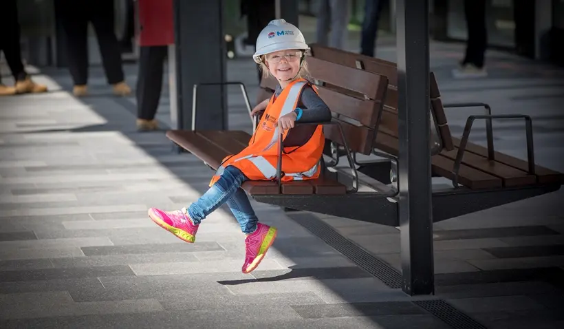 A child sitting on a bench wearing PPE vest, hardhat and glasses at Sydney Metro's Tallawong Station as part of the community day event.