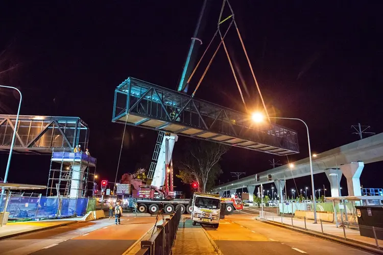 An on the ground view showing a crane lifting a segment of the pedestrian walkway into place at night at Sydney Metro's Kellyville Station.