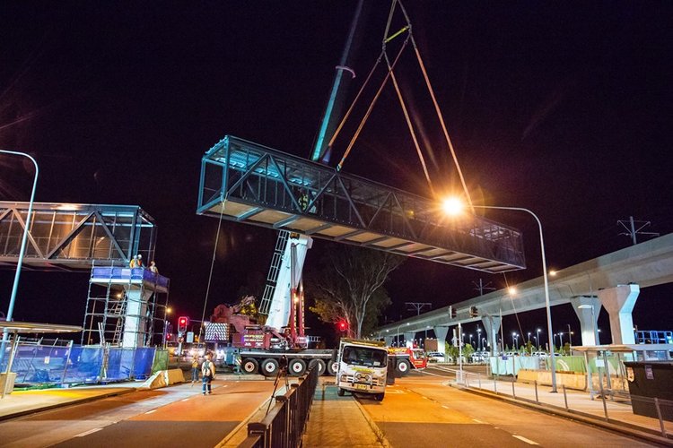 An on the ground view showing a crane lifting a segment of the pedestrian walkway  into place  at night at Sydney Metro's Kellyville Station.